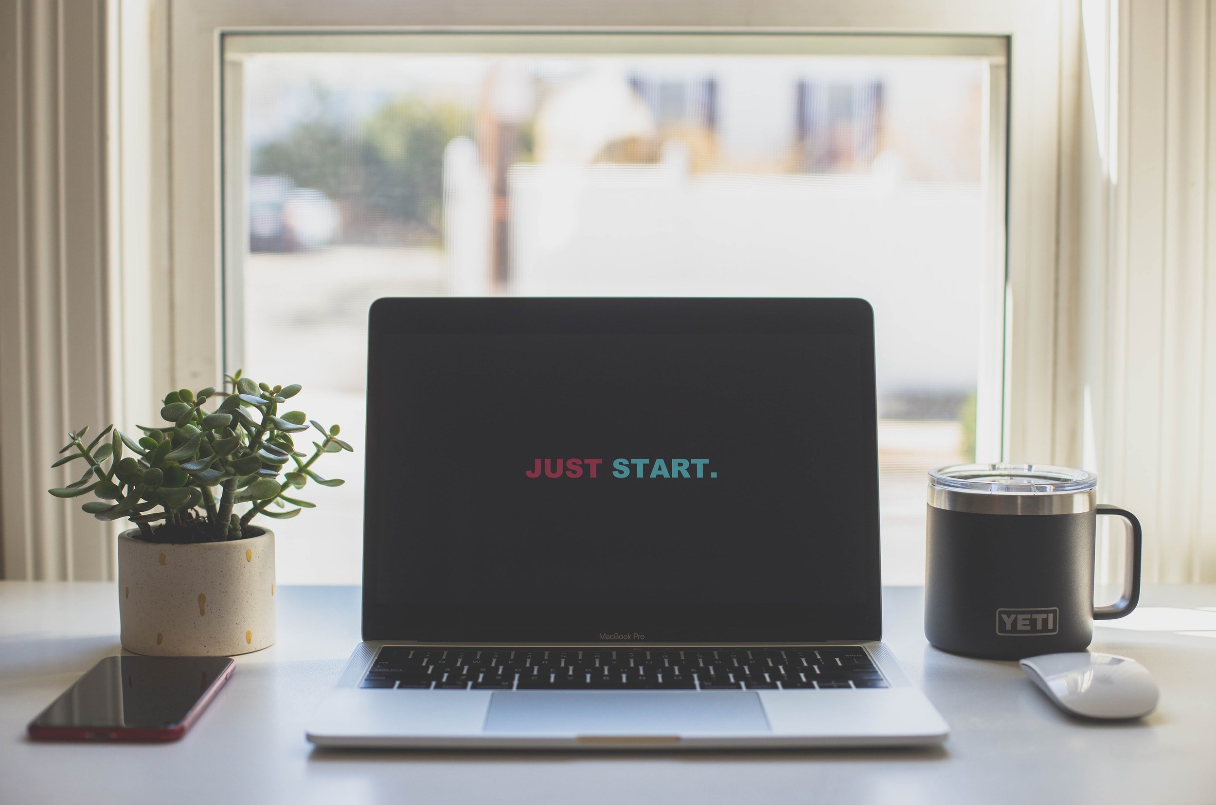 An Apple laptop in front of a window with a Yeti mug, an iPhone, and a magic mouse with the words "Just Start" on the screen 