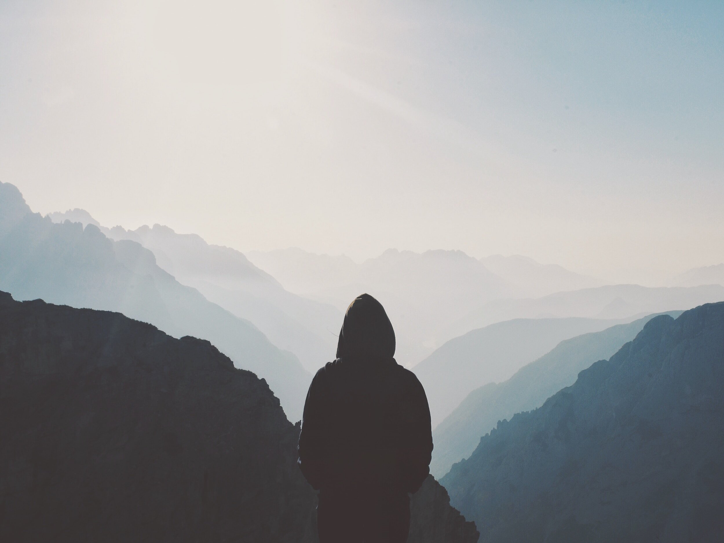 A person wearing a hoodie with their hands in their pockets, gazing at the sunny, misty mountains.