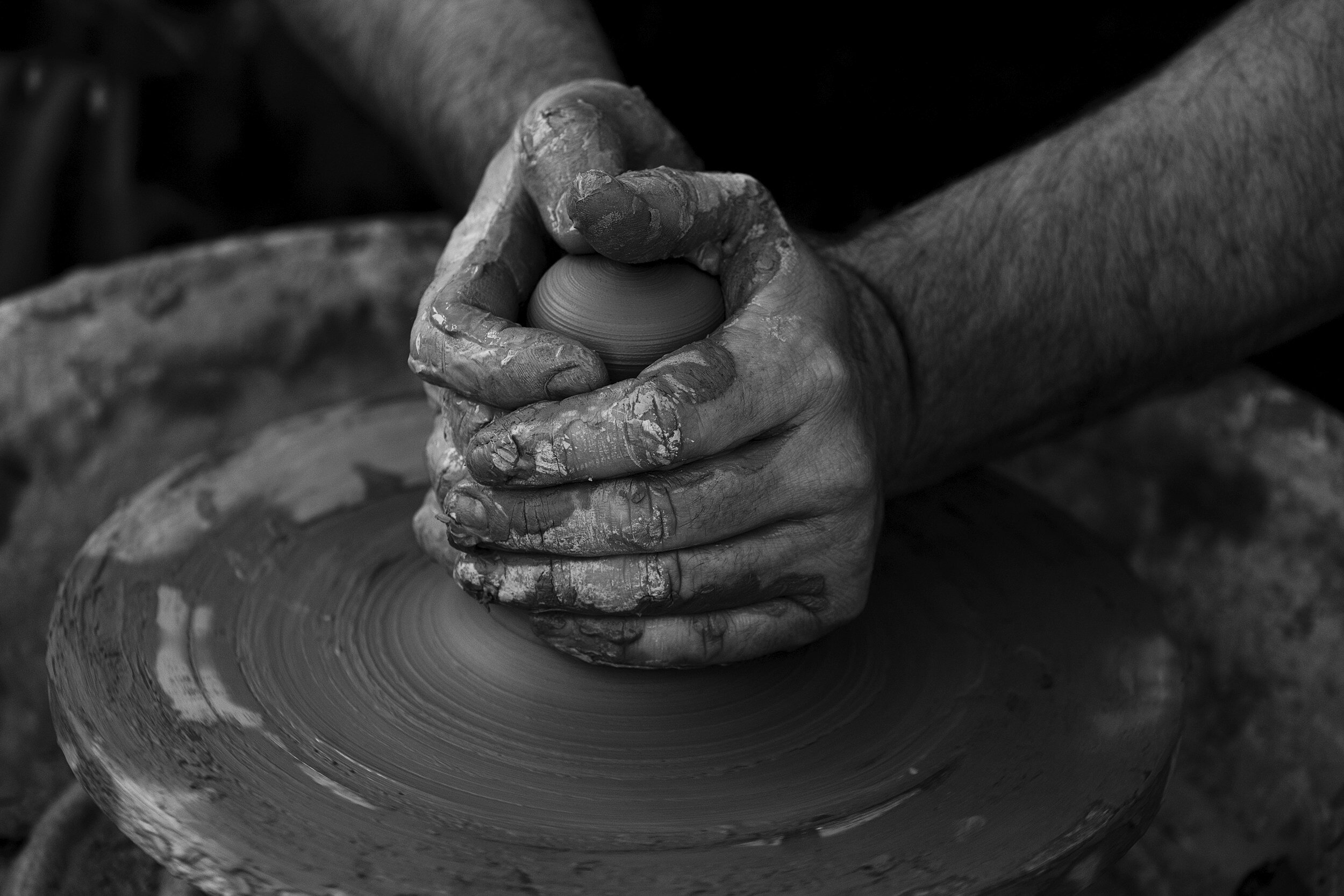 Black and white image of a potter's clay-covered hands shaping a vessel on a spinning pottery wheel.