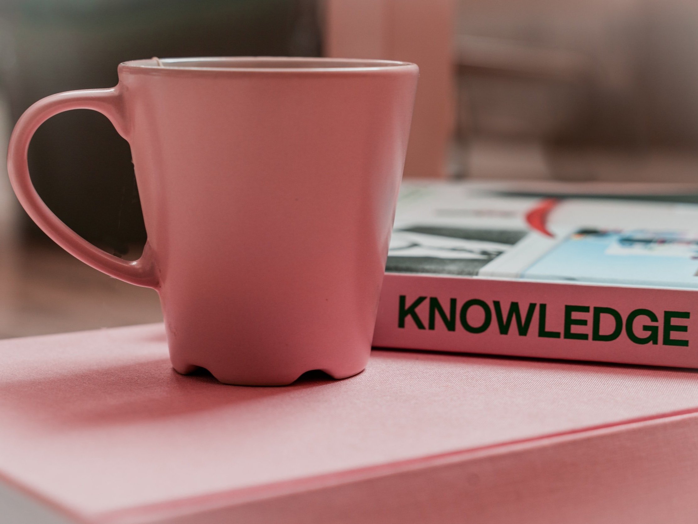 A pink coffee mug sits next to a pink book with the word knowledge written on the spine.