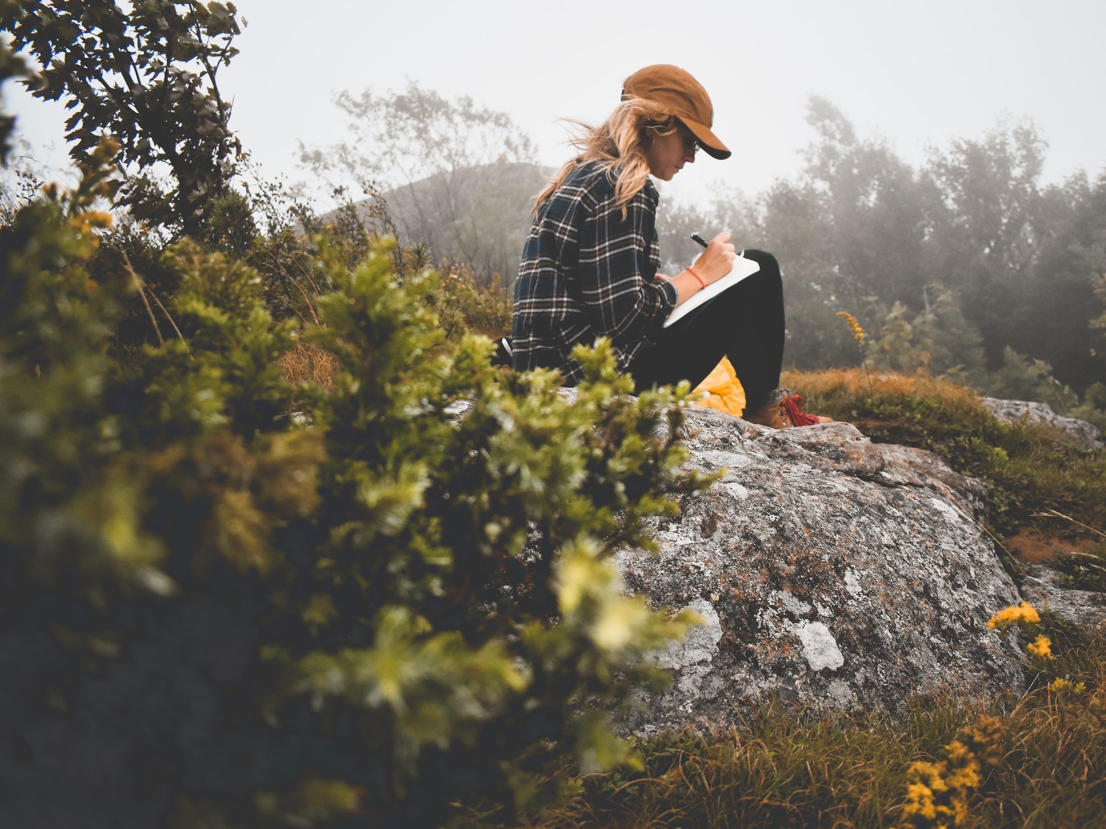 A woman writes while sitting on a rock in the countryside.