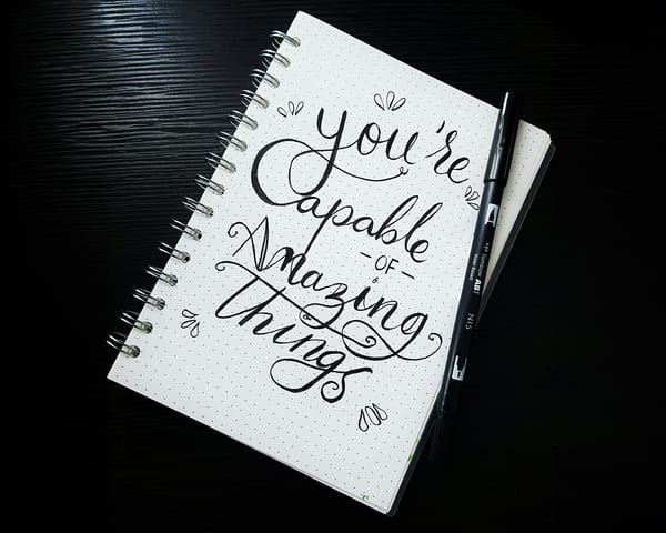 Notebook with inspirational quote "You're capable of amazing things" in calligraphy, alongside a pen.