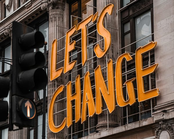 Neon sign reading "LET'S CHANGE" on a building, beside a traffic light, inspiring transformation in design.