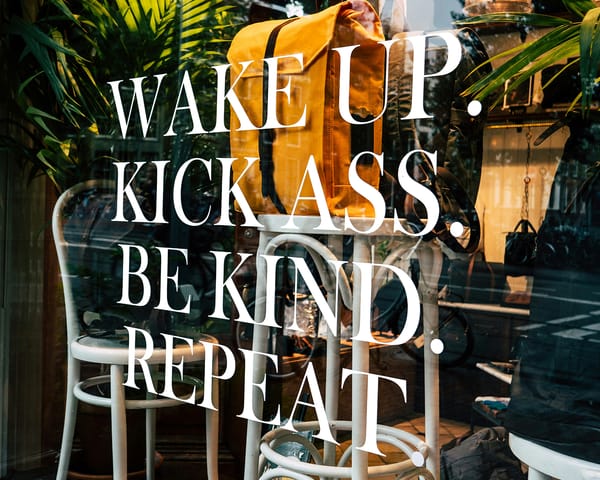 Inspirational quote "Wake up. Kick ass. Be kind. Repeat." over a window display with plants and chairs.