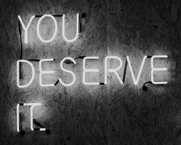 Neon sign "YOU DESERVE IT" on textured wooden background in black and white, sending an encouraging, motivational message.