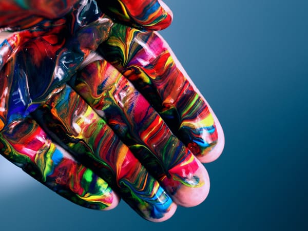 Open hand covered in vibrant, multi-colored paint - red, blue, yellow, green swirls and marbling on blurred blue background.