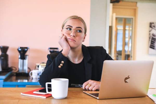 Person in glasses looking bored while sitting at a desk with a laptop, notebook, and coffee cup.