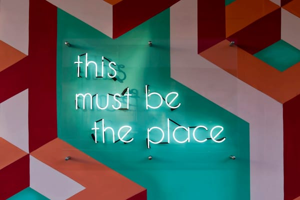 Neon sign saying "this must be the place" on a geometrically patterned wall, evoking a modern, creative vibe.