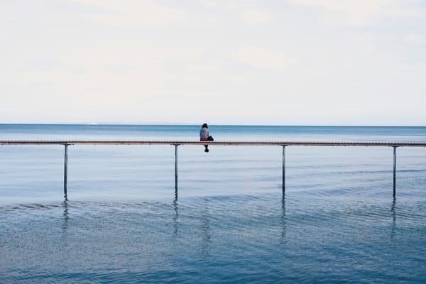 Person sits on serene pier over calm blue water under clear sky, conveying solitude and reflection.