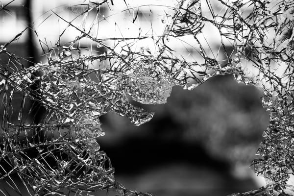 B&W photo of shattered glass pane, cracks forming web-like pattern, gaps revealing blurred background.