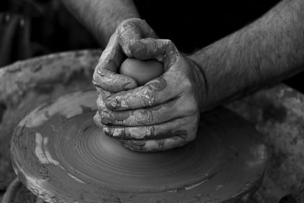 Black and white image of a potter's clay-covered hands shaping a vessel on a spinning pottery wheel.
